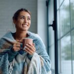 happy woman wrapped in a blanket drinking tea by the window