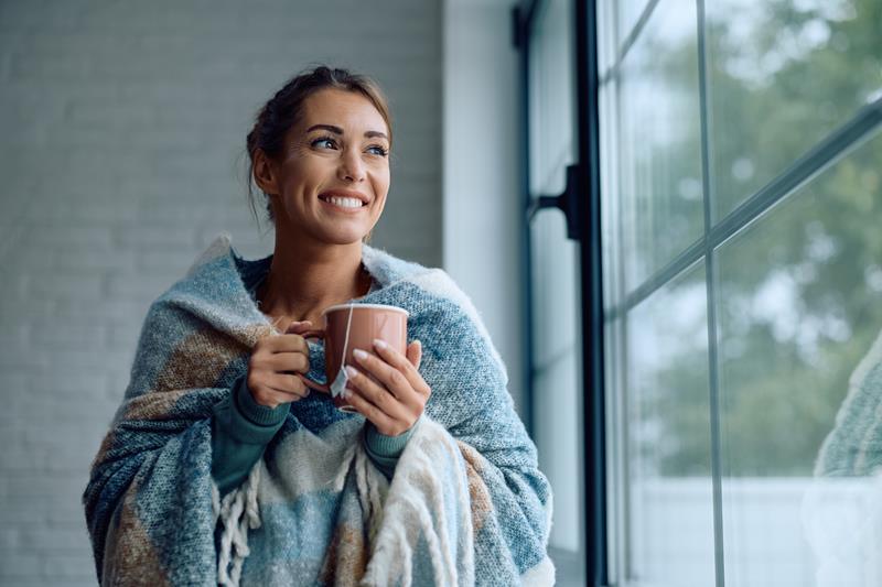 happy woman wrapped in a blanket drinking tea by the window